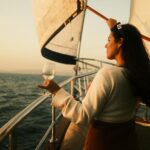 A woman stands on a sailboat, holding a glass of wine, enjoying the sunset view over the sea.