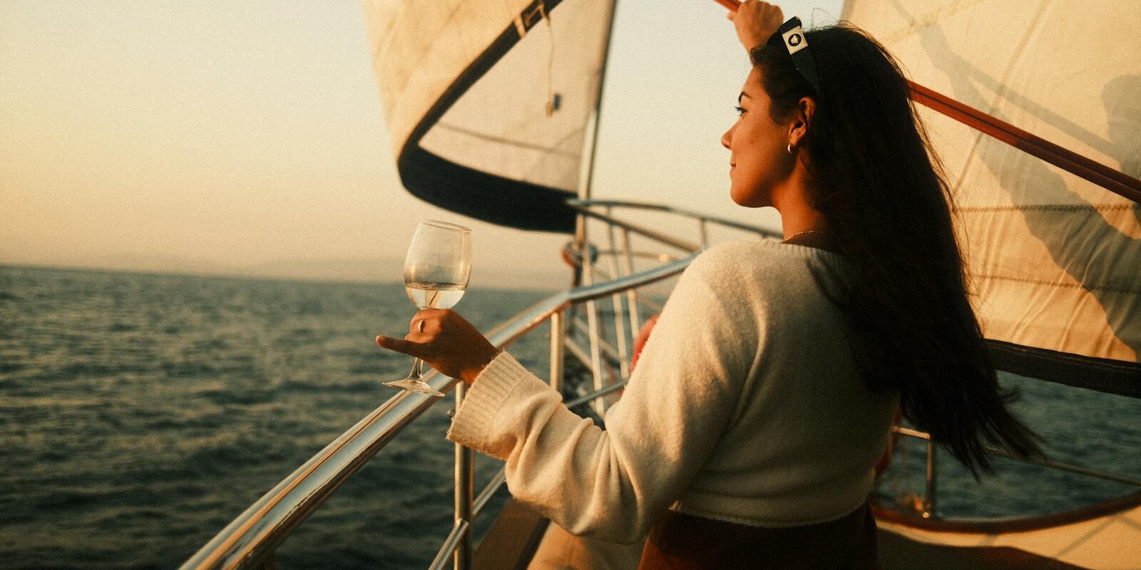 A woman stands on a sailboat, holding a glass of wine, enjoying the sunset view over the sea.