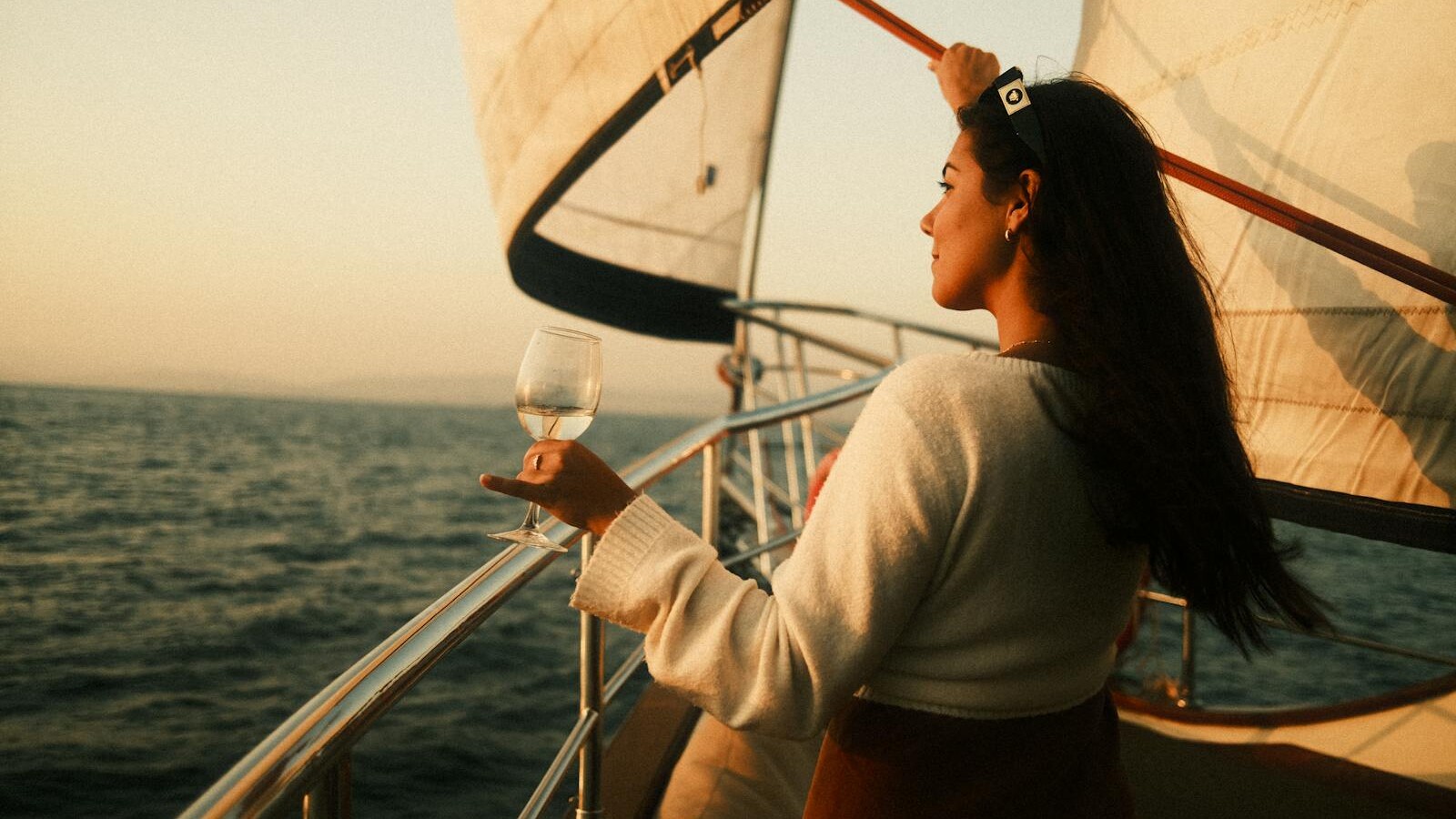 A woman stands on a sailboat, holding a glass of wine, enjoying the sunset view over the sea.