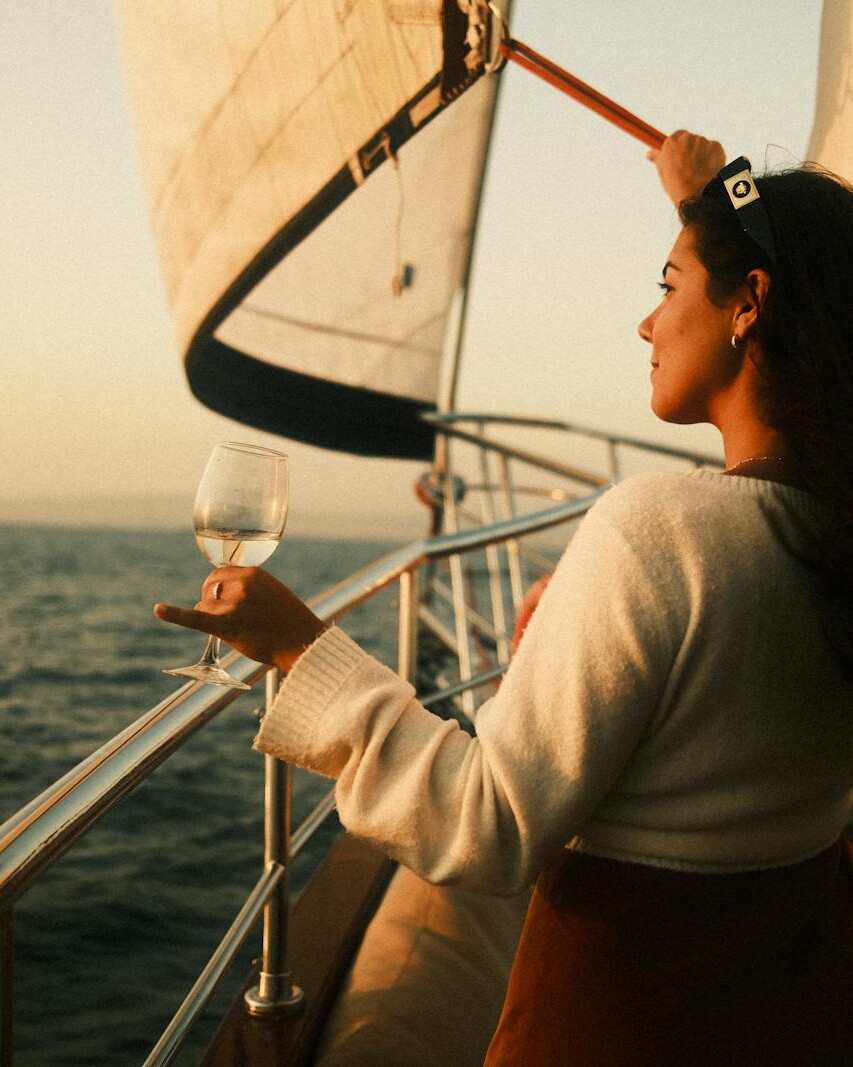 A woman stands on a sailboat, holding a glass of wine, enjoying the sunset view over the sea.