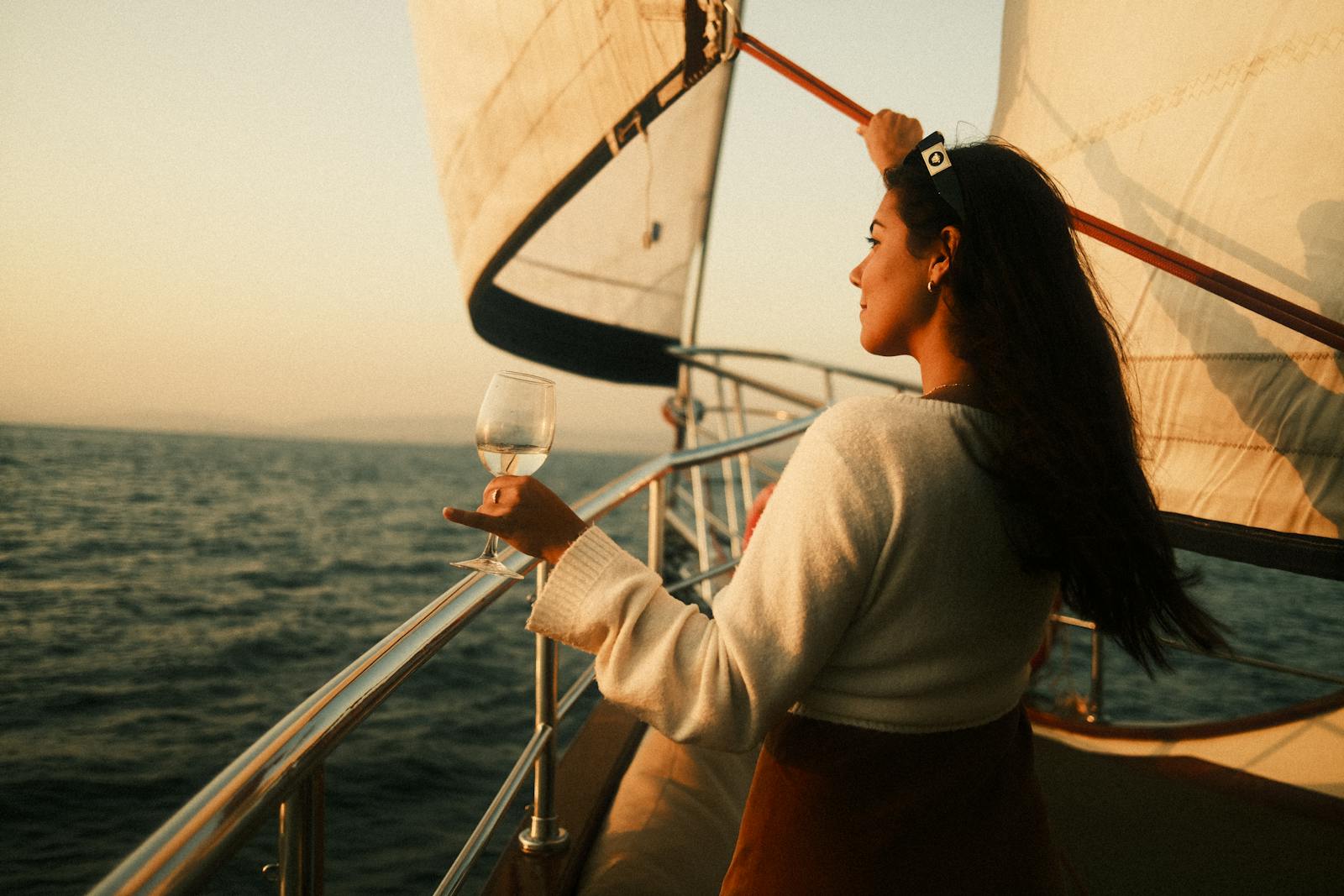 A woman stands on a sailboat, holding a glass of wine, enjoying the sunset view over the sea.